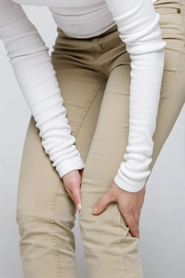 A woman wearing a white long-sleeve shirt and tan pants, clutching her knee, suffering from arthritis. Get arthritis treatment from Shelley Cole, M.D. of Healthy Success in Temple, Texas.