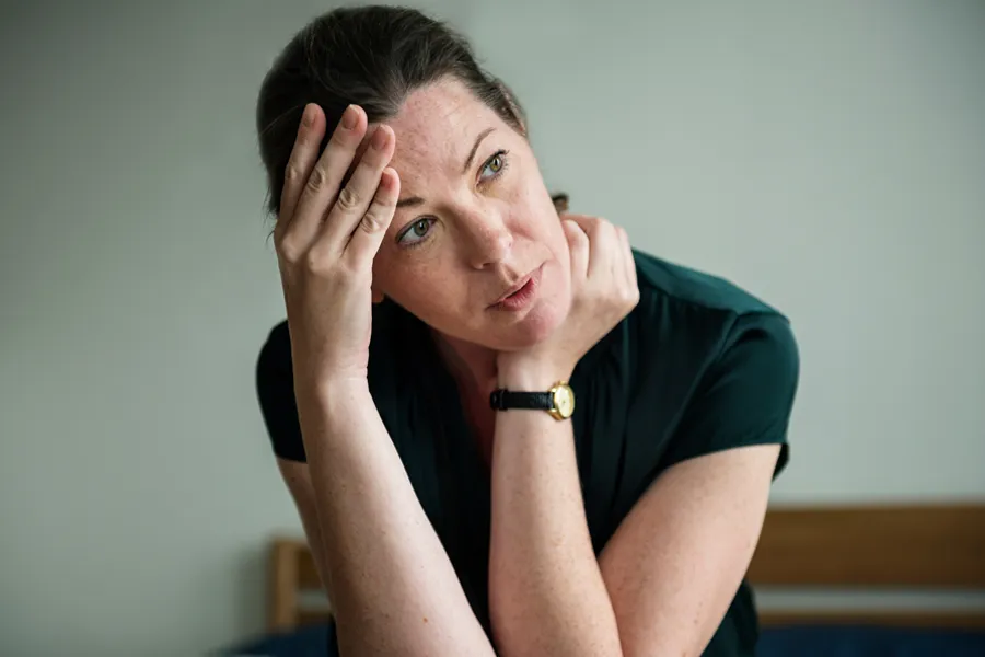 A woman in a dark blue dress sits on her bed holding her head in her hands with an exhausted expression. Get help for chronic fatigue syndrome from Shelley Cole, M.D. of Healthy Success in [AERA].