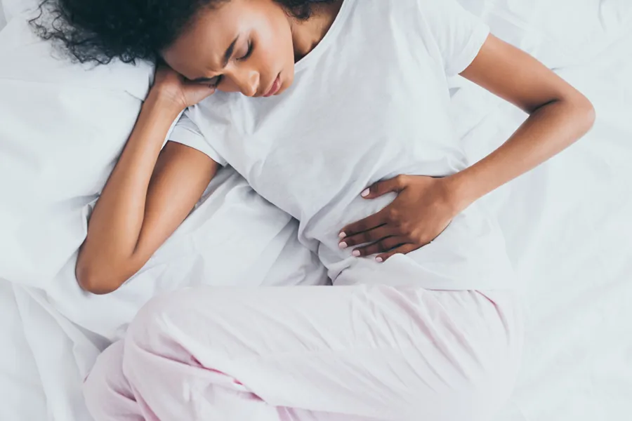 A dark-skinned woman in white clothes lays on her bed clutching her middle before getting treatment for Crohn's Disease from Shelley Cole, M.D. of Healthy Success in Temple, Texas.