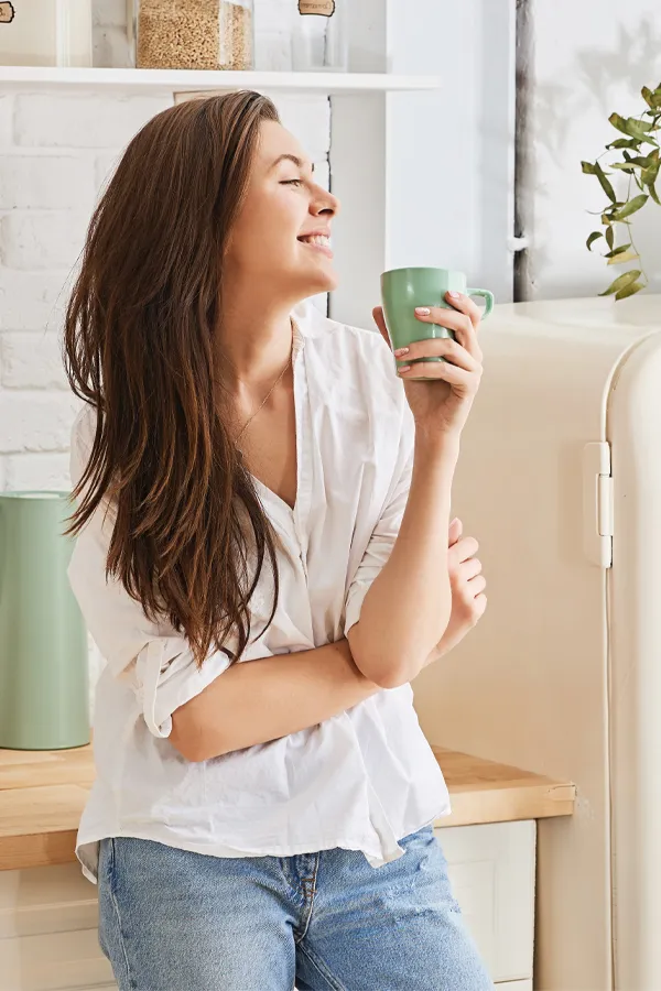 A woman with a white blouse an jeans drinking tea in her kitchen, smiling after getting treatment for Crohn's Disease from Shelley Cole, M.D. of Healthy Success in Temple, Texas.