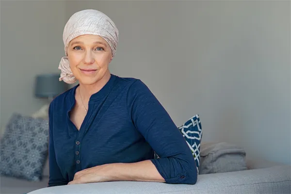 A woman wearing a blue shirt with a white scarf tied around her head sitting on a couch and smiling at the camera, representing sensitive functional oncology treatment from Shelley Cole, M.D. of Healthy Success in Temple, Texas.