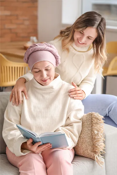 A woman with a scarf tied around her head sits on a couch reading while another woman sits behind her with her hands on her shoulders warmly, representing sensitive functional oncology treatment from Shelley Cole, M.D. of Healthy Success in Temple, Texas.