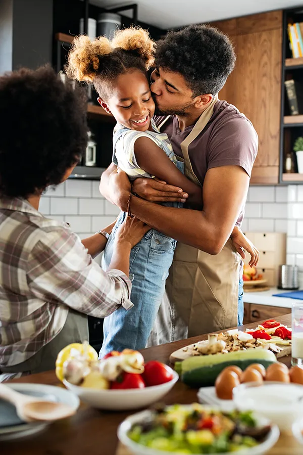 A man holds his daughter and kisses her cheek as the family makes a healthy dinner in the kitchen. Get help for digestive issues and gut health from Shelley Cole, M.D. of Healthy Success in Temple, Texas.