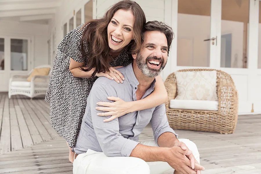 A middle-aged couple on a wooden porch with wicker furniture; the woman is playfully hugging the man from behind. They are benefiting from the health and wellness services from Shelley Cole, M.D. of Healthy Success in Temple, Texas.