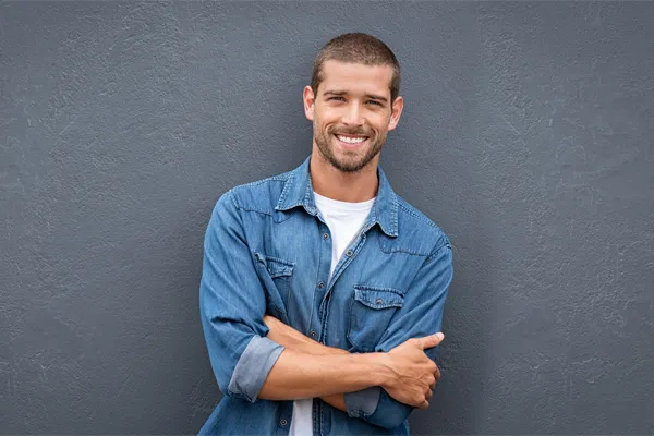 A man in a denim shirt stands smiling against a gray-blue wall, pleased with his testosterone hormone therapy from Shelley Cole, M.D. of Healthy Success in Temple, Texas.