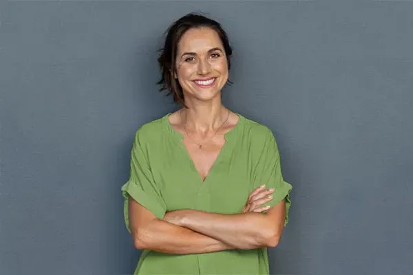 A woman in a green shirt stands smiling against a gray-blue wall, pleased with her hormone therapy from Shelley Cole, M.D. of Healthy Success in Temple, Texas.