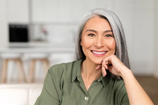 An asian woman with long gray hair and a green shirt sitting and smiling on her couch, happy with her menopause care from [NAME} in Temple, Texas.