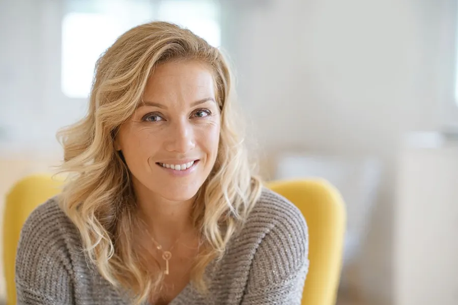 A blonde woman wearing a a gray sweater sits inside in a yellow chair, smiling and happy with her perimenopause treatment from Shelley Cole, M.D. of Healthy Success in Temple, Texas.