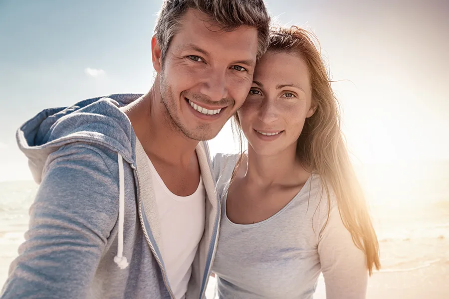 A middle-aged couple stands on the beach with the sun behind them. Get treatment for medical skin conditions from Shelley Cole, M.D. of Healthy Success in Temple, Texas.